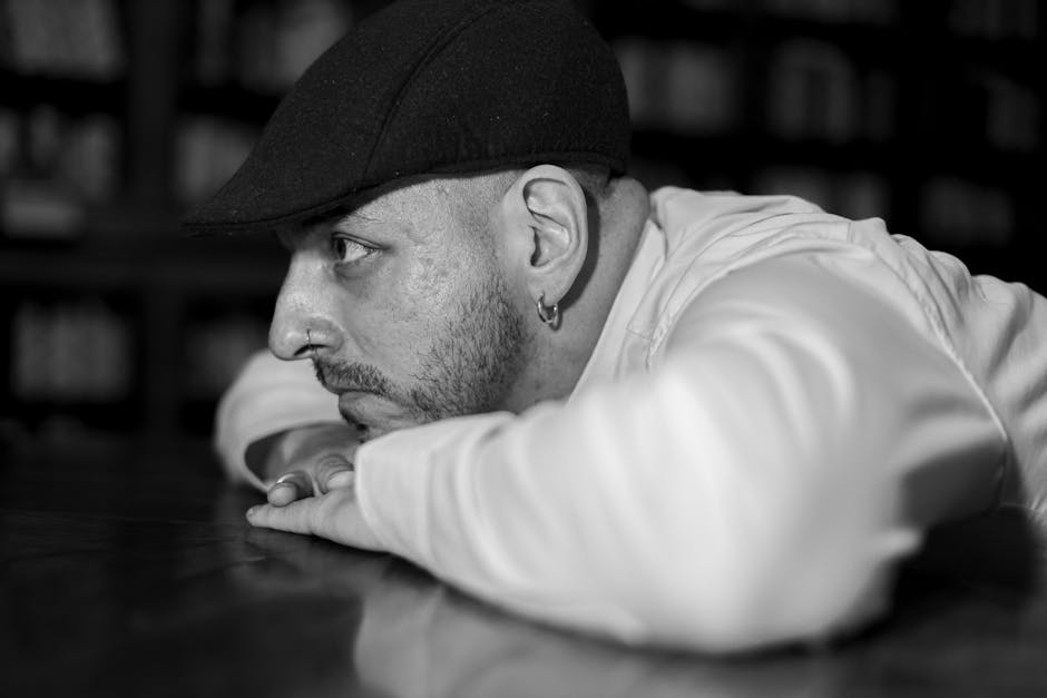 Siyah beyaz fotoğrafta, kütüphanedeki bir masanın üzerinde duran düşünceli bir adam görülüyor.