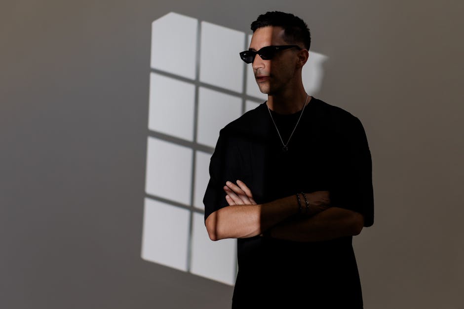 Kapalı alanda, sanatsal ışık ve gölge fonunda duran güneş gözlüklü bir adamın portresi.