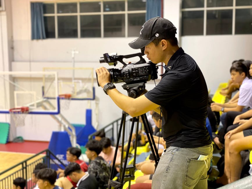Kameraman, arka planda seyircilerin olduğu bir iç mekan basketbol maçını kaydediyor.