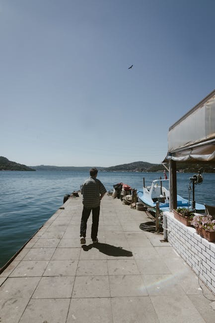 Bir adam, güneşli bir İstanbul iskelesinde tekneler ve berrak gökyüzü eşliğinde huzurlu bir yürüyüşün tadını çıkarıyor.