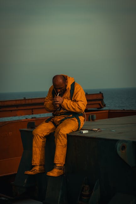 Gündüz vakti çekilen bu görüntüde, sarı yağmurluk giymiş yalnız bir işçi, denizdeki bir gemide otururken sigara yakıyor.