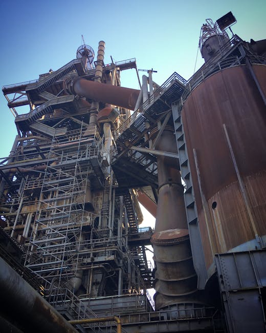 Açık gökyüzü altında, karmaşık boru tesisatı ve iskele sistemine sahip büyük bir endüstriyel çelik yapının görüntüsü.