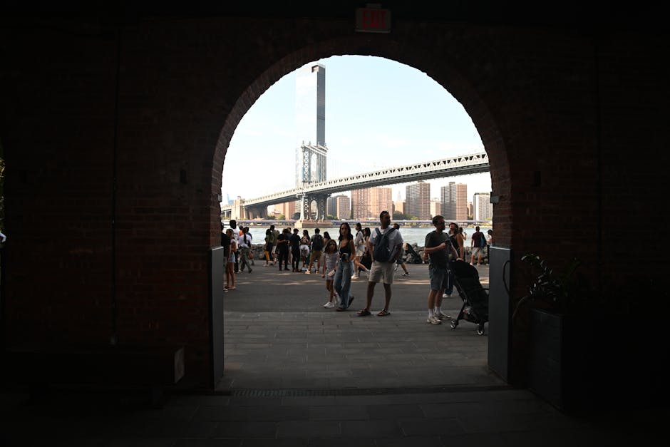 Güneşli bir günde, tuğla bir kemerle çerçevelenmiş Manhattan Köprüsü'nün yakınında insanlar toplanıyor.