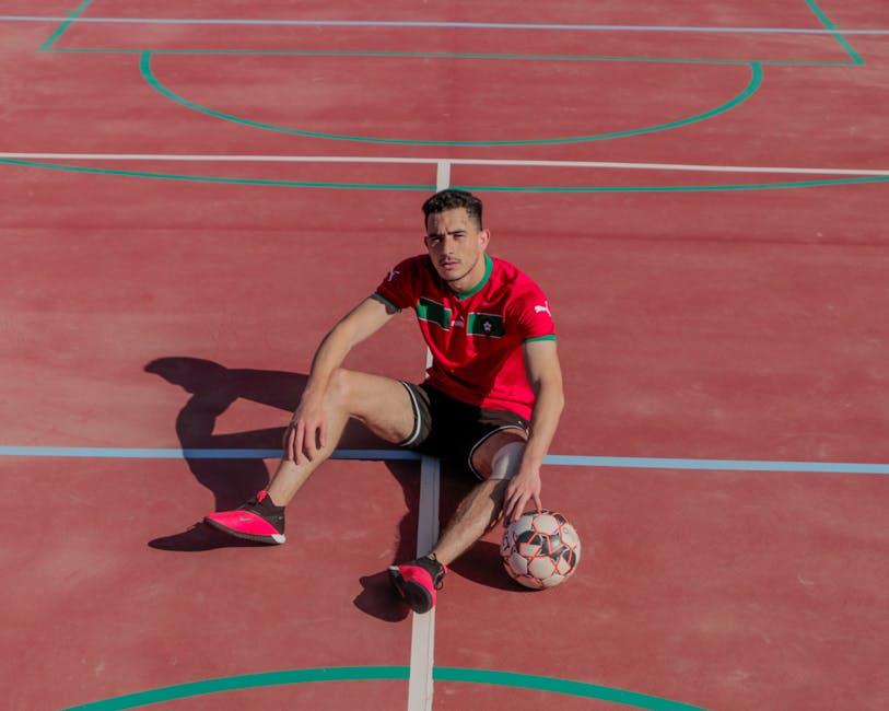 Spor kıyafetli atletik adam, elinde futbol topuyla kırmızı bir açık hava sahasında oturuyor.