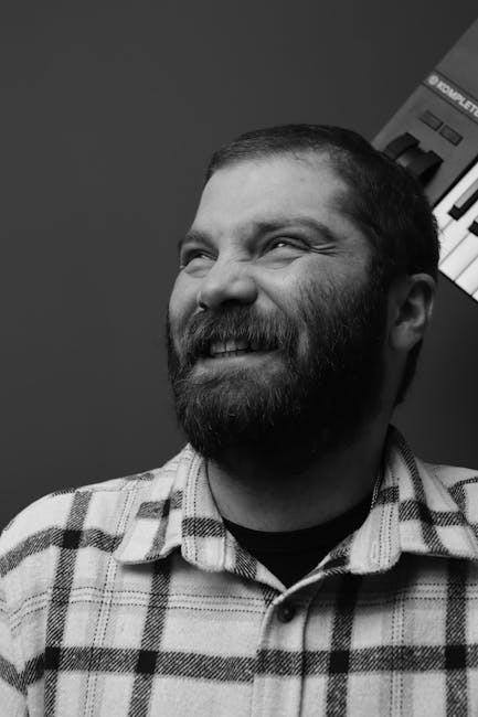 Arka planda müzik klavyesi bulunan, gülümseyen sakallı bir adamın siyah beyaz portresi.