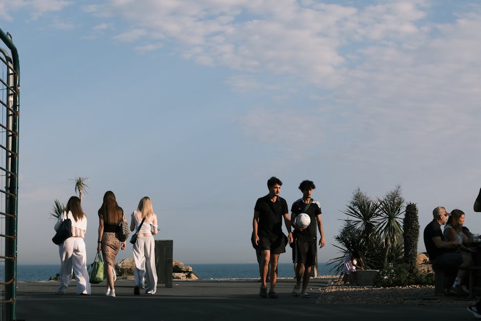 Deniz kenarında yürüyüş yapan ve futbol oynayan iki gencin yanında keyif yapan insanlar.